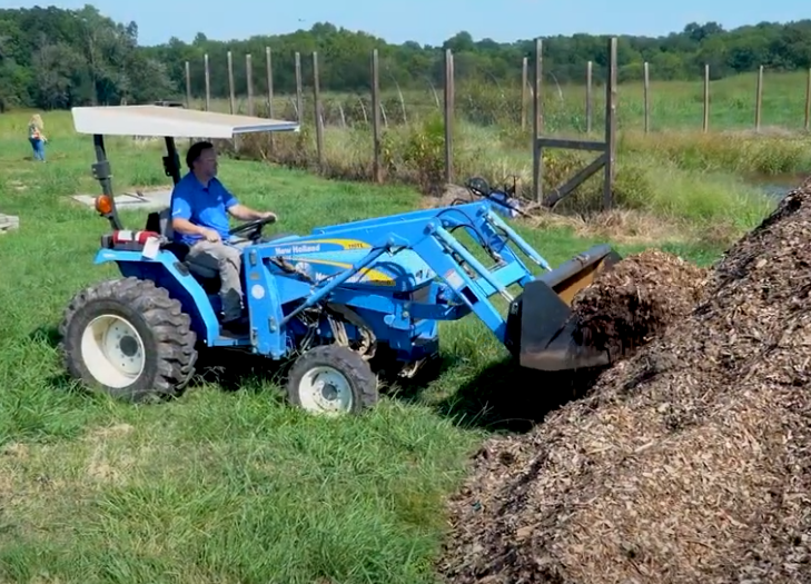 A student operates a blue tractor to move mulch at ACC’s farm site. The scene shows a hands-on agricultural learning environment with fields and fencing in the background.