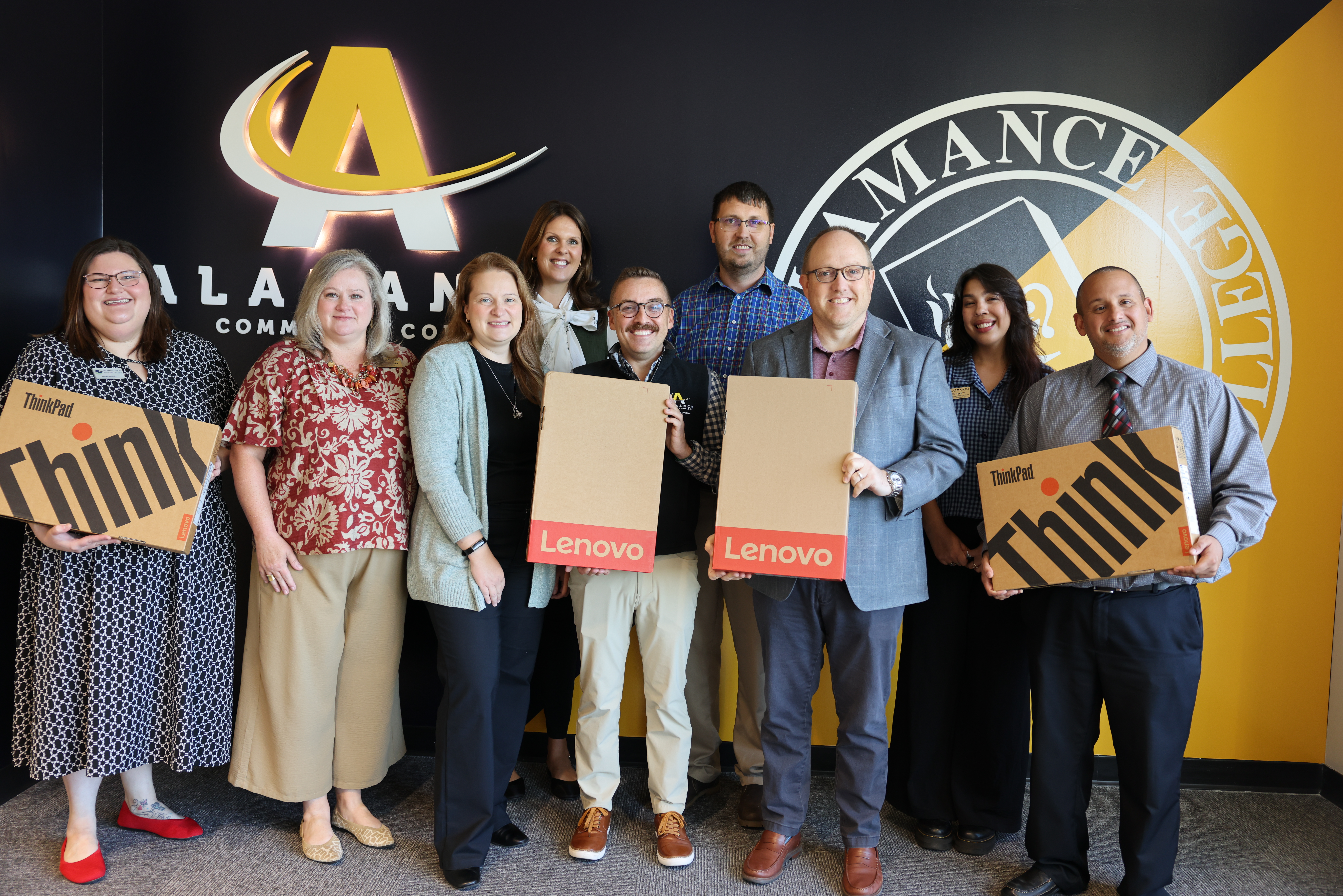 Nine people smile in front of the Alamance Community College logo, holding Lenovo ThinkPad laptop boxes.
