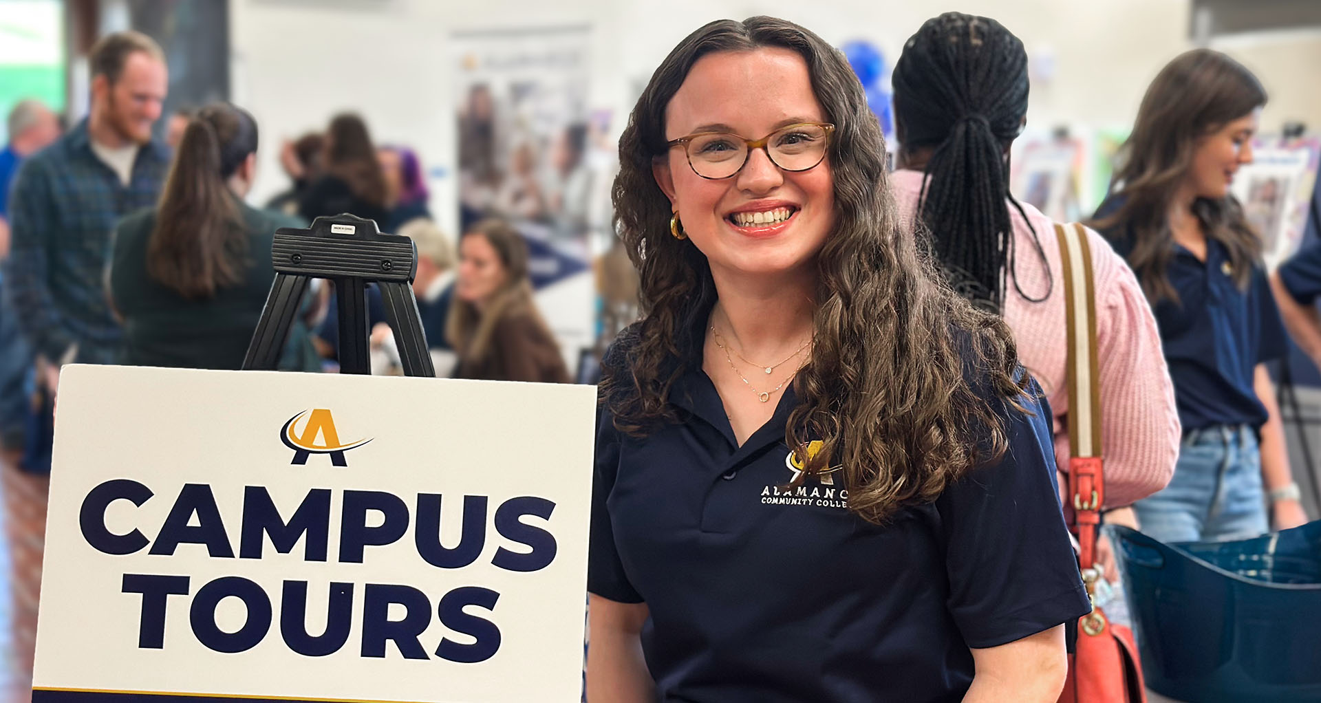 A smiling ACC representative stands beside a sign that reads 'Campus Tours' during a busy campus event. She is wearing an ACC-branded navy polo shirt, and the background is filled with attendees engaging with displays and staff.