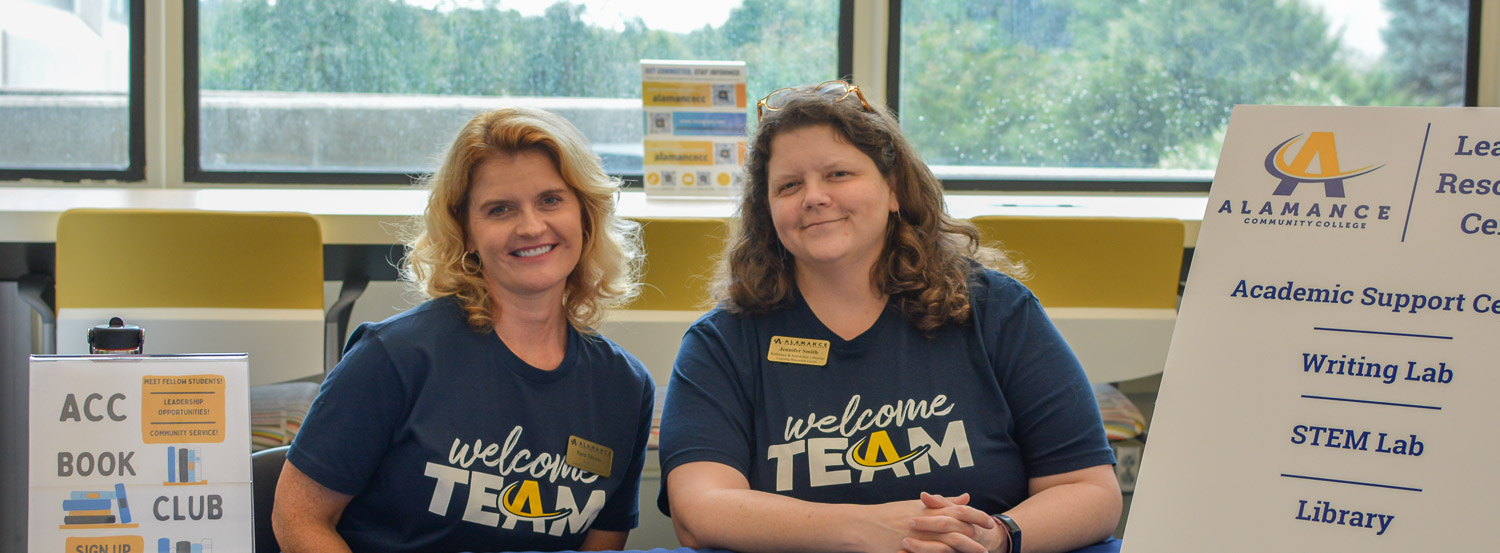Two Alamance Community College staff members seated at a welcome table promoting student support services, including the Academic Support Center, Writing Lab, STEM Lab, Library, and ACC Book Club.