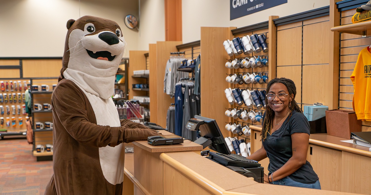 Bookstore Alamance Community College mascot Otter stands at the campus bookstore counter, interacting with a smiling staff member. The background features ACC merchandise, including apparel and drinkware."