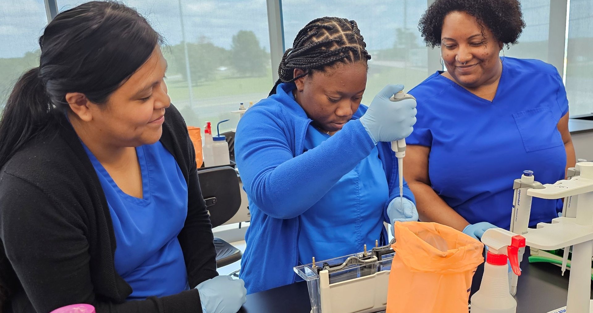 Three Alamance Community College Medical Laboratory Technology students wearing blue scrubs work together in a bright lab. One student uses a pipette while the others observe and support her. The workspace is filled with lab equipment and supplies, emphasizing hands-on experience and collaborative learning.