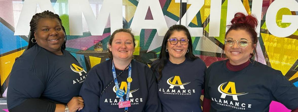 ACC Staff Four smiling Alamance Community College staff wearing ACC shirts stand in front of a colorful wall mural that reads 'YOU ARE AMAZING.' The group poses confidently, showcasing school spirit and community pride.