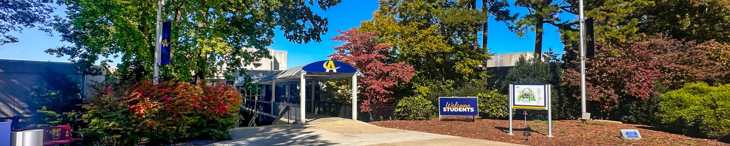 Main entrance to Alamance Community College on a bright, sunny day, featuring a covered walkway with the ACC logo, a 'Welcome Students' sign, and colorful trees and landscaping surrounding the path.