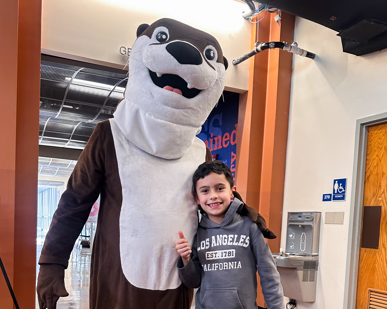 Young boy smiling and giving a thumbs up while posing with Alamance Community College’s otter mascot inside a campus building.