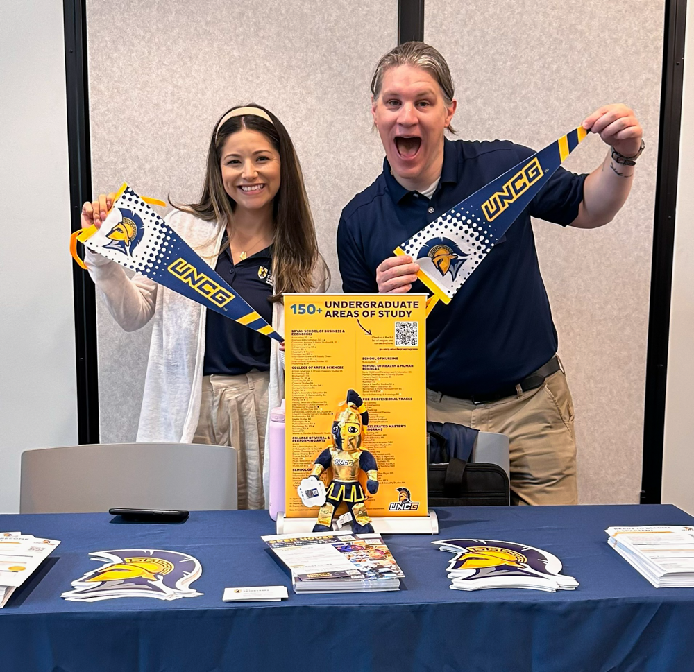 Two college representatives smiling and holding UNCG pennants behind a table with brochures and information about undergraduate programs.