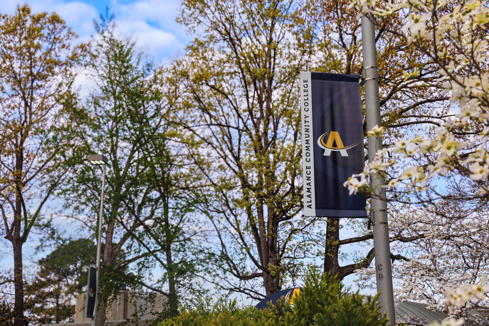 Alamance Community College banner hanging on a campus light pole surrounded by trees in bloom.