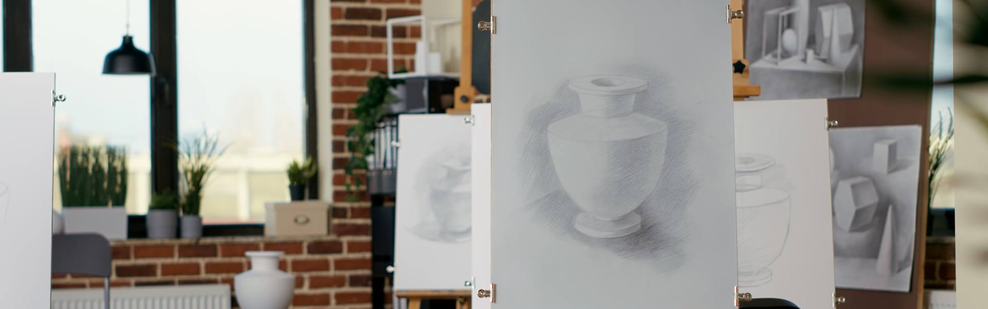 Art classroom with easels displaying pencil drawings of still-life objects, including a shaded vase, in a well-lit studio with brick walls and windows.